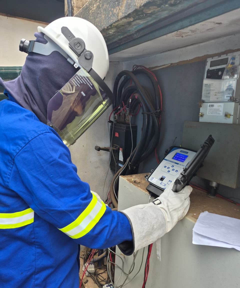 Técnico com capacete, balaclava, protetor facial e luvas isolantes fazendo medição com analisador portátil em quadro elétrico aberto